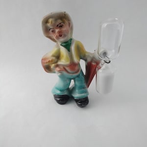 May include: A ceramic figurine of a person wearing a hat and holding an hourglass. The figurine is painted in shades of blue, yellow, red, and white.