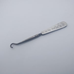 May include: A vintage silver-toned button hook with a curved hook end and a decorative handle. The handle features an engraved floral design. The tool is designed for fastening buttons on shoes, gloves, or clothing.