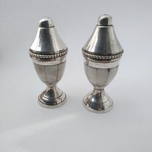 May include: Two silver salt and pepper shakers. Each shaker has a conical top and a base with a pedestal foot. The body of each shaker is decorated with a mother-of-pearl inlay. A beaded band encircles the body.