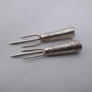May include: Two silver-toned metal hors d'oeuvre forks featuring three slender tines and a conical handle. The handles are decorated with a repeating engraved pattern of small circular swirls and leaf-like motifs across the entire surface.