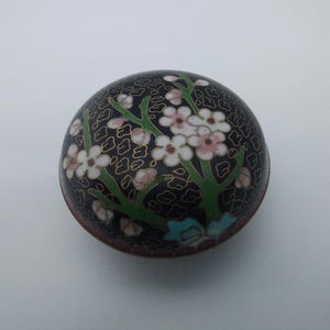 May include: A round, black decorative box with a floral design. The box features white and pink flowers with green stems against a black background. The flowers are accented with gold detailing. The box is likely made of metal.
