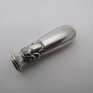Könnte beinhalten: Ein silberfarbener Wachssiegelstempel mit einem ovalen Griff und aufwendigen floralen Details. Der Griff ist glatt mit einem Perlenrand, und die Basis weist ein dekoratives Design auf. Der Stempel wird wahrscheinlich zum Versiegeln von Dokumenten verwendet.