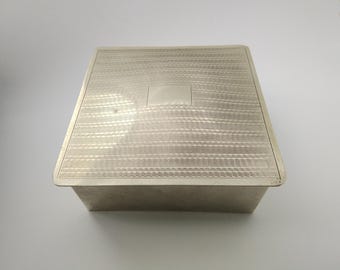 May include: A silver-colored rectangular box with a textured lid. The lid features a repeating wave-like pattern and a small, rectangular, unadorned area in the center. The box is likely used for storage or display.