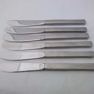 May include: A collection of five stainless steel butter knives. Each knife features a broad, curved blade and a textured handle. The blades are polished, reflecting light, and the handles have a rough, hammered finish. The knives are laid out horizontally.