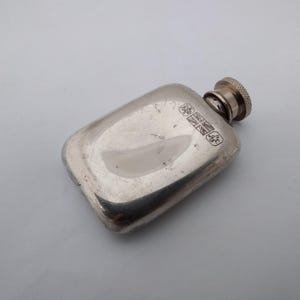 May include: A rectangular silver-toned metal flask with rounded corners and a textured screw-top cap. The surface features a series of small, stamped hallmarks near the neck, including a Union Jack flag symbol and other distinct markings.