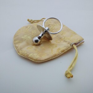 May include: A shiny silver-coloured dummy with a round handle and a circular shield, resting on a small, pale yellow drawstring pouch featuring a subtle, repeating floral pattern woven into the fabric of the bag.