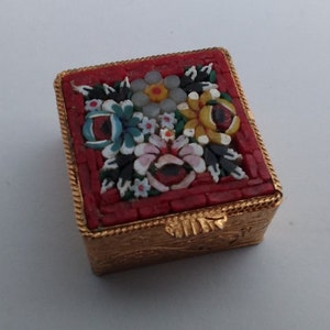 May include: A small, square, red and gold mosaic trinket box with a floral design. The box is decorated with a variety of colourful flowers, including red, yellow, blue, and white.