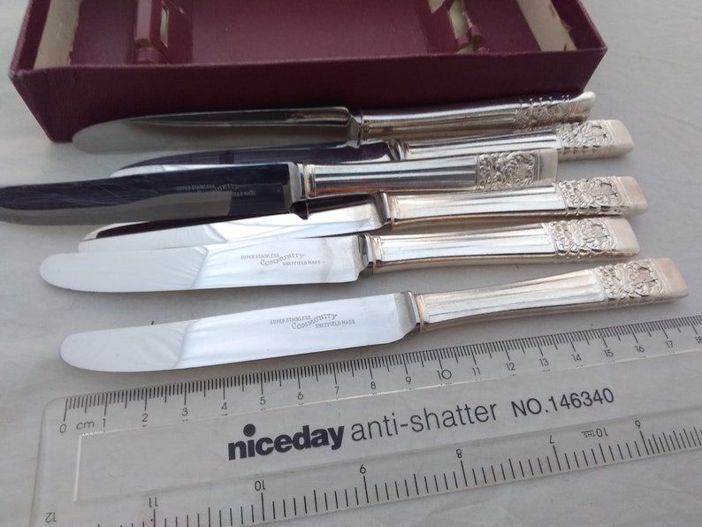 Puede incluir: Juego de seis cuchillos de mantequilla de plata con mangos ornamentados. Los cuchillos est&aacute;n dispuestos sobre una superficie blanca con una regla para la escala. Los cuchillos est&aacute;n marcados con "Super Stainless" y "Coopers Sheffield" en las hojas.