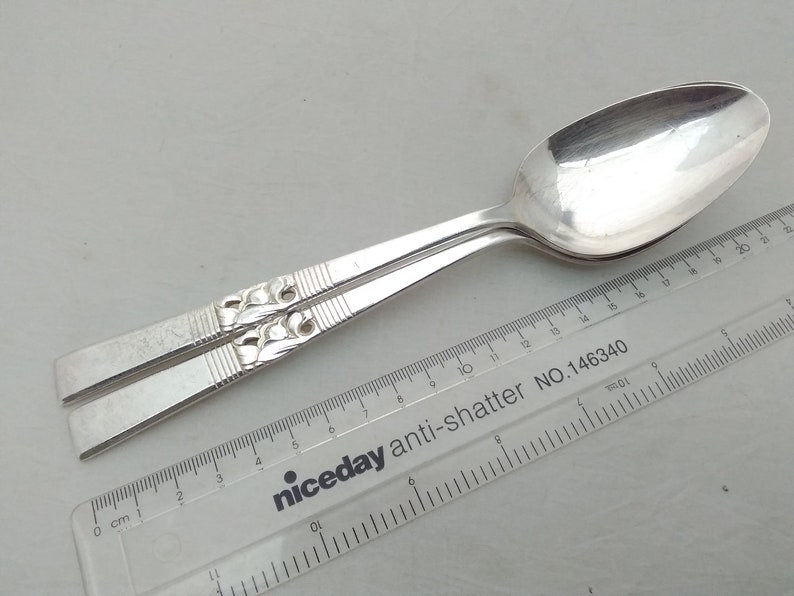 May include: Two silver spoons with an ornate design on the handle. The spoons are laid out on a white surface with a ruler that reads "niceday anti-shatter NO.146340".