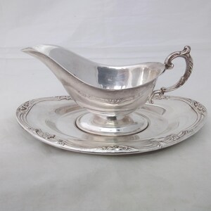 May include: A silver-toned gravy boat with a curved handle and a matching oval underplate. The gravy boat features a pedestal base and a smooth, polished finish. The underplate has a decorative embossed border with intricate scrollwork details.
