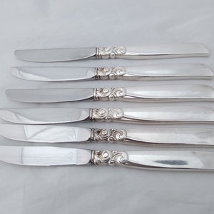 May include: Set of six silver butter knives with ornate handles. The knives have a polished finish and are arranged in a row.