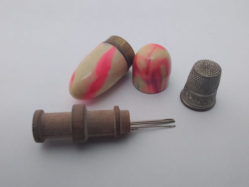 K&ouml;nnte beinhalten: Ein Vintage-N&auml;hset mit einem h&ouml;lzernen Nadelf&auml;der, einem Metall-Fingerhut und zwei rosa-wei&szlig; gestreiften Nadelk&auml;sten.