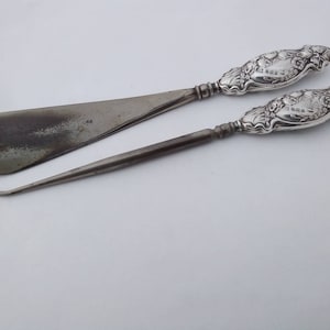 May include: A set of two silver shoe horns with ornate floral designs on the handles. The handles are marked with the text "1883".