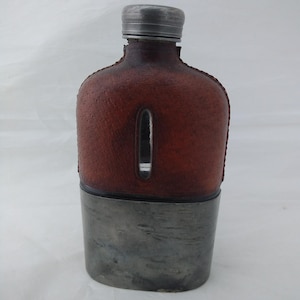 May include: A flask featuring a brown textured leather wrap on the upper portion with a vertical viewing window, a metallic screw-top cap, and a smooth, brushed silver-toned metal base. The container has a rounded rectangular shape.