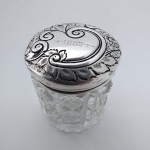 May include: A vintage silver-toned jar with a clear, faceted glass base and a decorative lid. The lid is engraved with floral patterns and the year "1903". The jar is likely a container for small items or trinkets.