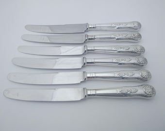 May include: Six silver knives with ornate handles. The blades are smooth and reflective, and the handles feature a detailed floral design. The knives are arranged in a row, showcasing their elegant design, perfect for a formal dining setting.