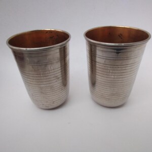 Puede incluir: Dos vasos de color plateado con interior color cobre. Los vasos tienen forma cilíndrica con líneas grabadas horizontales. Los vasos están sobre un fondo blanco.