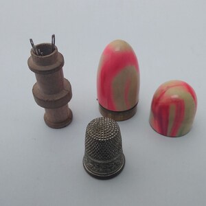 K&ouml;nnte beinhalten: Ein Set aus vier Vintage-N&auml;hwerkzeugen, darunter ein h&ouml;lzerner Fingerhut-Halter, zwei rosa-wei&szlig; gestreifte Fingerh&uuml;te und ein silberner Metallfingerhut.