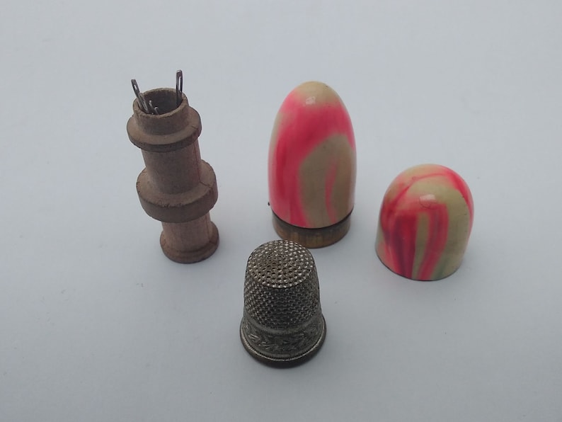 K&ouml;nnte beinhalten: Ein Set aus vier Vintage-N&auml;hwerkzeugen, darunter ein h&ouml;lzerner Fingerhut-Halter, zwei rosa-wei&szlig; gestreifte Fingerh&uuml;te und ein silberner Metallfingerhut.