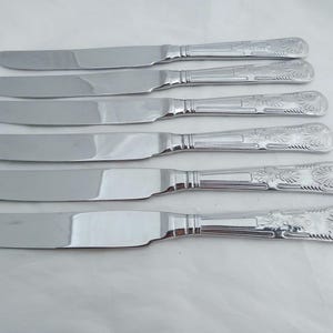 May include: A collection of six silver knives. Each knife features a long, flat blade and a decorative handle with an ornate floral design. The knives are displayed in a row on a white surface.