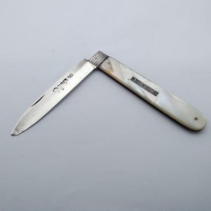 May include: A partially open pocket knife with a silver blade and a white handle. The handle has a mother-of-pearl appearance with a small decorative metal accent. The blade has text etched on it. The knife is set against a gray background.