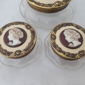 May include: Three glass jars with gold lids and white cameo portraits of women. The lids have a decorative floral design.