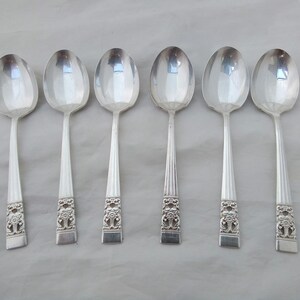 May include: Six silver spoons with ornate handles. The spoons are arranged in a row on a white surface.