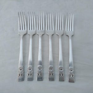 May include: A set of six silver forks. Each fork has multiple tines and a decorative handle with an ornate design. The handles feature a detailed pattern, possibly of trees or foliage. The forks are arranged in a row on a neutral surface.