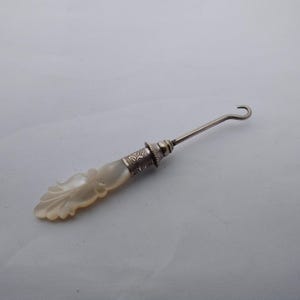 May include: A button hook featuring a carved mother-of-pearl handle with a leaf-like design. The handle connects to a slender metal shaft that terminates in a curved hook. A decorative silver-toned metal collar with an engraved floral pattern sits between the handle and the shaft.