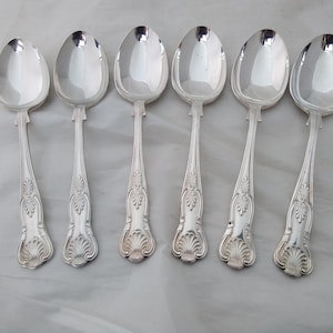 May include: Six silver spoons with oval bowls and ornate, detailed handles. The spoons are arranged in a row, reflecting light. The handles feature a decorative, stylized leaf and shell design, adding a touch of elegance to the silverware.