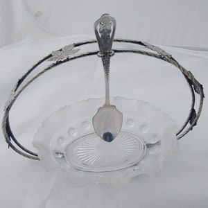 May include: A silver serving set with a glass bowl and a spoon. The bowl has a scalloped edge and a clear glass base. The spoon has a decorative handle with a floral design. The set is on a white background.