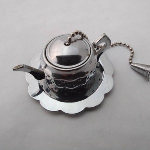 May include: A silver-coloured tea infuser in the shape of a teapot, sitting on a scalloped saucer. It has a chain with a bell-shaped end. The teapot infuser is designed for steeping loose leaf tea.