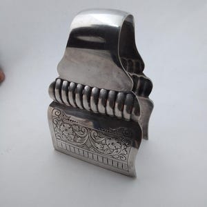 May include: A silver-toned metal object with a curved handle and a decorative floral design. The object has a ribbed section and a detailed engraved pattern. The piece is likely a vintage or antique item.