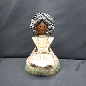 Op de afbeelding: Een keramische pop met een bruin gezicht en zwart haar. De pop draagt een witte jurk met een groene en bruine basis.