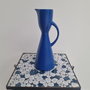 Könnte beinhalten: Eine blaue Keramikkanne mit breitem Boden und Henkel. Die Kanne steht auf einer weißen und blauen gefliesten Oberfläche.
