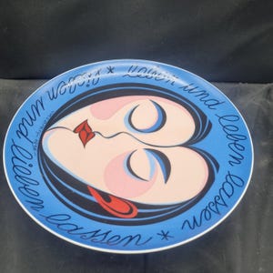 May include: A decorative plate with a blue rim and a central design featuring a stylized face. The face has black hair, pink cheeks, and a red lip. The plate has the text "lieben und leben lassen" in a cursive font.