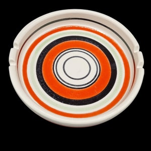 May include: A round, ceramic ashtray with a white rim and a concentric circle design in orange, black, green, and white. The ashtray has three small notches for resting cigarettes. The design is centered on a white base.
