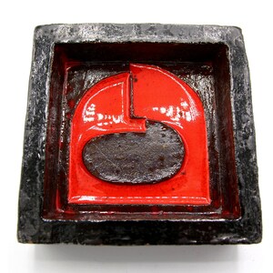 Könnte beinhalten: Ein roter und schwarzer Aschenbecher aus Keramik mit geometrischem Design. Der Aschenbecher hat eine quadratische Form mit einem erhöhten Rand und einer vertieften Mitte.