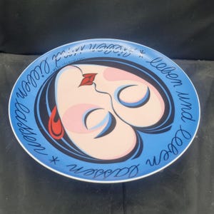 May include: A decorative plate with a blue rim and a central design featuring a stylized face with red lips and a black outline. The plate has the text "leben und lieben lassen" in a cursive font around the edge.