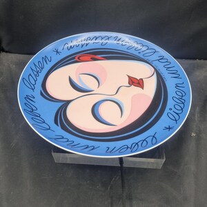 May include: A decorative plate with a blue rim and a central design featuring a stylized face with closed eyes and a red lip. The face is surrounded by a blue background with the words "Leben und Lieben lassen" in a circular pattern.