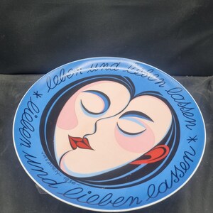 May include: Decorative plate with a blue border and a central design featuring two stylized faces in profile, with closed eyes and red lips. The plate has the text "lieben und lieben lassen" and "vasarely-1936".