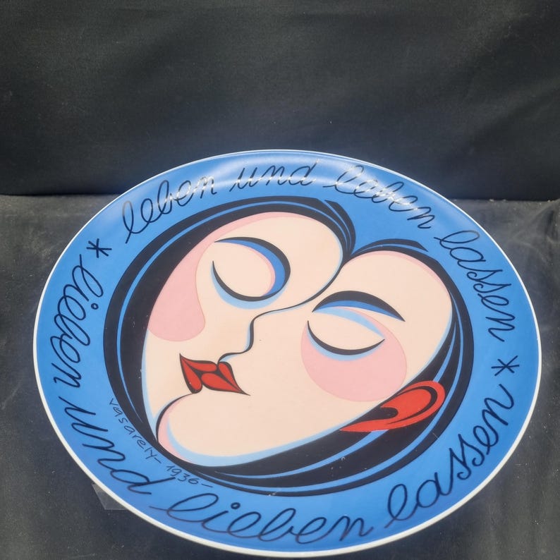 May include: Decorative plate with a blue border and a central design featuring two stylized faces in profile, with closed eyes and red lips. The plate has the text "lieben und lieben lassen" and "vasarely-1936".