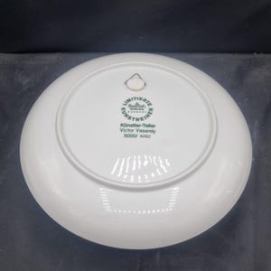 May include: A white porcelain plate with a circular design. The back of the plate features a green and white emblem with German text, including "LIMITIERTE KUNSTREIHE" and "Victor Vasarely". The plate also has a small metal hanger.