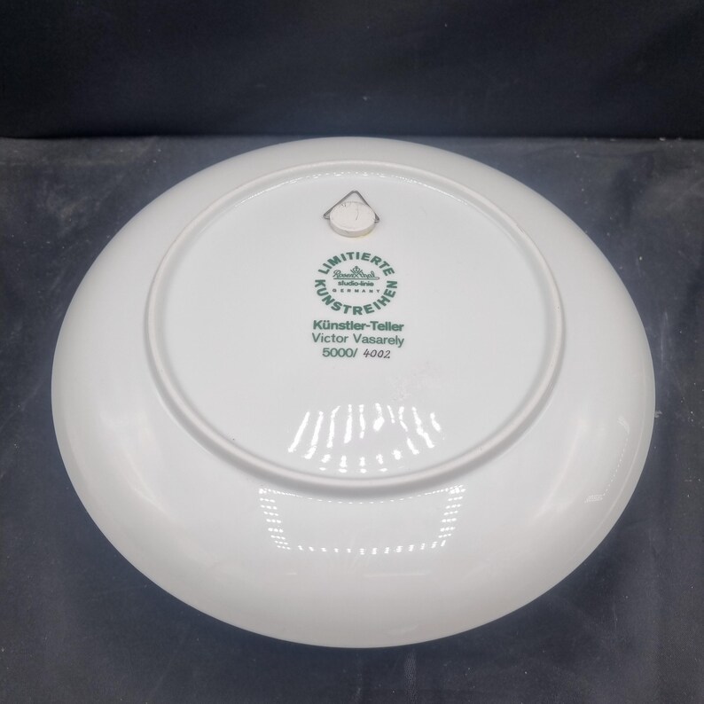May include: A white porcelain plate with a circular design. The back of the plate features a green and white emblem with German text, including "LIMITIERTE KUNSTREIHE" and "Victor Vasarely". The plate also has a small metal hanger.