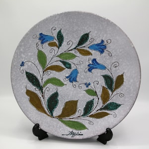 May include: A white ceramic plate featuring a blue and green floral design. The plate has a textured surface and is signed "Baylis" in the bottom right corner.