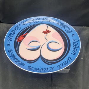 May include: A decorative plate with a blue rim and a central design featuring two faces kissing, forming a heart shape. The faces have pink skin, red lips, and black hair. The plate has the text "Leben und lieben lassen" in a cursive font.