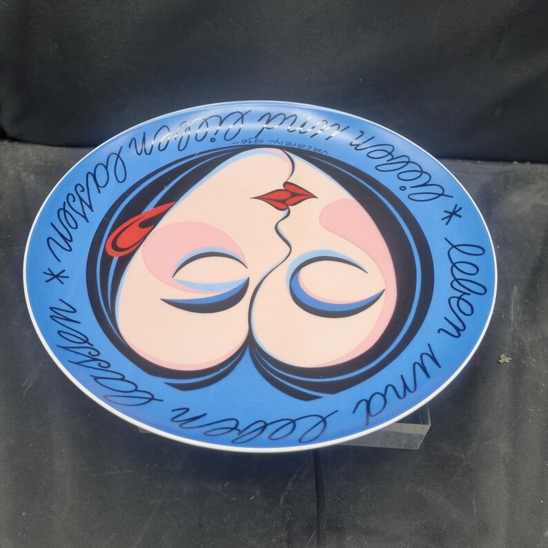 May include: A decorative plate with a blue rim and a central design featuring two faces kissing, forming a heart shape. The faces have pink skin, red lips, and black hair. The plate has the text "Leben und lieben lassen" in a cursive font.