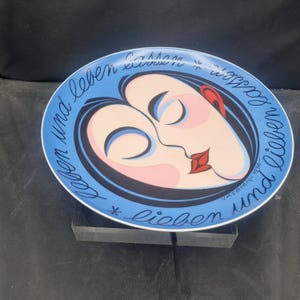 May include: A decorative plate with a blue rim and a central design featuring two stylized faces in profile, with red lips and ears. The plate has text in black cursive around the edge, and is signed "vasarely-1936".