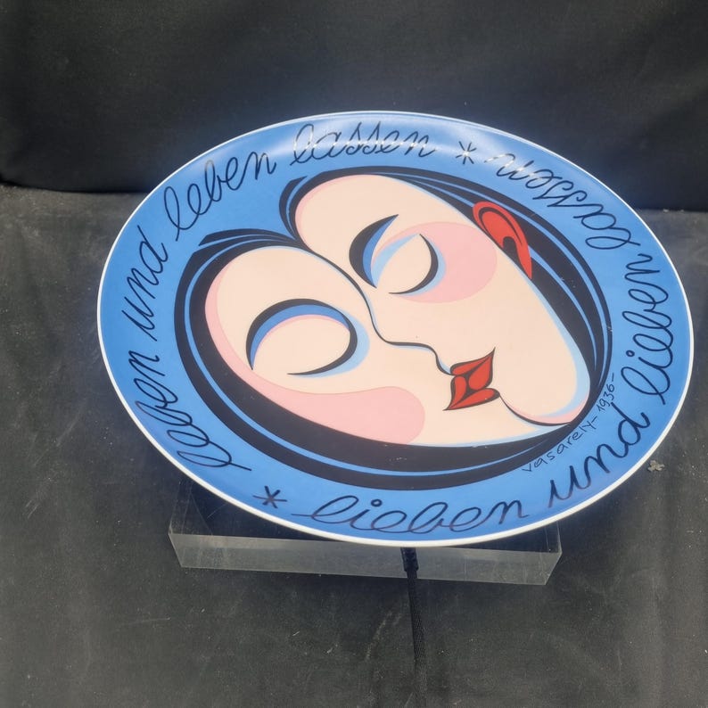 May include: A decorative plate with a blue rim and a central design featuring two stylized faces in profile, with red lips and ears. The plate has text in black cursive around the edge, and is signed "vasarely-1936".