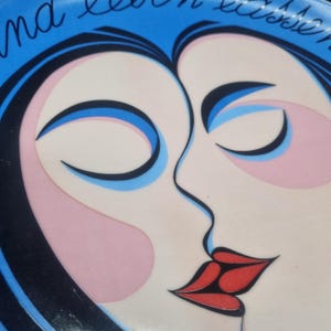 May include: A close-up of a blue and white decorative plate featuring a stylized illustration of two faces kissing. The faces have closed eyes, pink cheeks, and red lips. The plate has text in cursive.